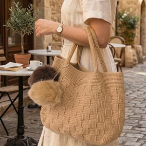 Könnte beinhalten: Eine beigefarbene Häkeltasche mit einem Webmuster, getragen von einer Person. Die Tasche hat zwei flauschige Pompons. Der Text "VISUALMINTS" befindet sich oben links. Der Text "INSTANT PDF DOWNLOAD - CROCHET PATTERN" befindet sich unten.