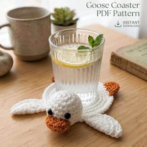 Peut inclure: Un dessous de verre en crochet blanc en forme d'oie, avec un bec et des pieds orange, supporte un verre d'eau avec du citron et de la menthe. Le dessous de verre est posé sur une surface en bois. Le texte sur l'image indique "VisualMints Goose Coaster PDF Pattern".