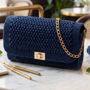 Könnte beinhalten: Eine dunkelblaue gehäkelte Handtasche mit strukturiertem Maschenmuster, einem goldfarbenen Metallkettengriff und einem rechteckigen goldfarbenen Drehverschluss. Zwei goldfarbene Häkelnadeln liegen im Vordergrund.