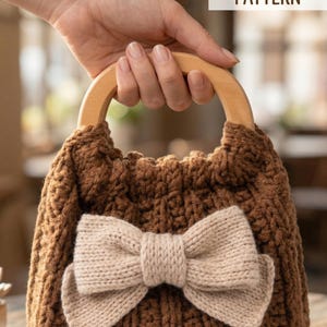 Könnte beinhalten: Eine braune gehäkelte Handtasche mit einer hellbeigen gestrickten Schleife auf der Vorderseite und einem abgerundeten Holzgriff. Der Text PDF CROCHET PATTERN ist in der oberen rechten Ecke des Bildes sichtbar.