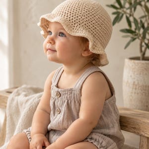 May include: A beige crochet bucket hat with a ruffled brim worn with a light beige sleeveless romper featuring two small brown buttons on the front bodice and elasticized leg openings, creating a soft and neutral textured outfit ensemble.