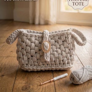 Könnte beinhalten: Eine hellgraue, grob strukturierte Tragetasche mit goldenem Verschluss. Die Tasche hat zwei Griffe und besteht aus einem dicken, gewebten Material. Ein Häkelhaken und ein Knäuel Garn liegen neben der Tasche. Der Text auf dem Bild lautet "CROCHET PATTERN PDF DOWNLOAD" und "CHUNKY TEXTURE TOTE".