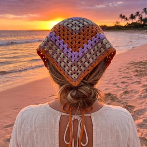 Könnte beinhalten: Ein dreieckiges Häkelkopftuch mit Granny-Square-Muster in den Farben Orange, Lavendel und Creme. Das Kopftuch verfügt über lange, gehäkelte orangefarbene Bänder mit Quasten an den Enden, die am Hinterkopf zusammengebunden werden können.