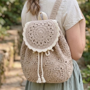 Könnte beinhalten: Beigefarbener Häkelrucksack mit kreisförmiger Klappe und Kordelzug. Die Klappe hat ein detailliertes, spitzenartiges Design mit cremefarbenem Besatz. Der Rucksack hat verstellbare Träger und einen oberen Griff.