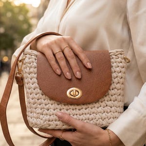 Könnte beinhalten: Eine beige gehäkelte Handtasche mit einer braunen Kunstlederklappe, einem goldfarbenen Metalldrehverschluss und einem passenden braunen Schulterriemen. Die Tasche hat eine strukturierte, gewebte Optik und enthält ein kostenloses Tragetaschenmuster.