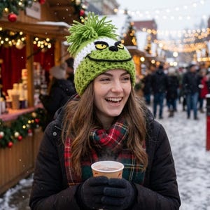 Könnte beinhalten: Eine lindgrüne Häkelmütze mit einem großen Bommel oben und einem Charaktergesicht mit gelben Augen, schwarzen Augenbrauen und einem breiten, schwarz gestickten Lächeln. Die Mütze hat ein weißes Band über dem Gesicht und einen rot-grünen Schal.