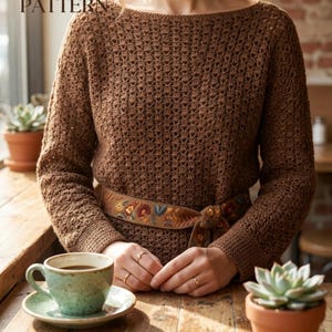 Könnte beinhalten: Ein brauner Häkelpullover mit Bootsausschnitt und langen Ärmeln. Der Pullover wird mit einem braunen Gürtel mit floraler Stickerei getragen. Der Text "CROCHET PDF PATTERN" und "BOHO LACE SWEATER" ist sichtbar. Eine Tasse Kaffee und eine Sukkulente sind ebenfalls zu sehen.