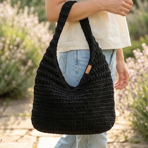Könnte beinhalten: Eine schwarze gehäkelte Tragetasche mit einem einzelnen Schulterriemen und einem kleinen rechteckigen braunen Lederetikett an der Seite. Die Tasche hat ein strukturiertes Rippenmuster und enthält den Text Bonus Free Tote Bag Pattern.