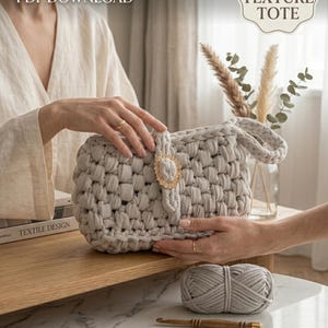Könnte beinhalten: Eine hellgraue, grob strukturierte Tragetasche mit goldenem Verschluss, hergestellt aus dickem Garn. Das Bild zeigt einen Wollknäuel, eine Häkelnadel und den Text "CROCHET PATTERN PDF DOWNLOAD" und "CHUNKY TEXTURE TOTE".