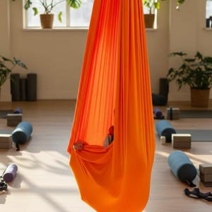 May include: Bright orange aerial yoga hammock suspended in a sunlit studio. The fabric is draped, creating a cocoon-like shape. Yoga mats, blocks, and bolsters are visible on the wooden floor, along with potted plants near a window.