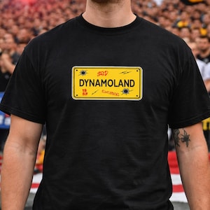 May include: Black t-shirt featuring a yellow license plate design with the text "GOD DYNAMOLAND 19 53 ELBLORENZ". The shirt is a classic crew neck style.
