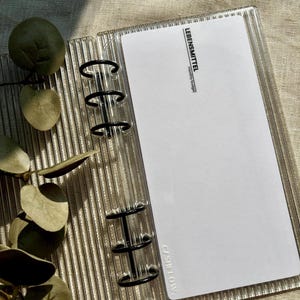Könnte beinhalten: Transparenter, strukturierter Acryl-Ordner mit schwarzen Metallringen, der ein weißes Blatt Papier enthält. Der Text "LEBENSMITTEL monthly budget" ist auf das Papier gedruckt. Der Ordner ist geöffnet.