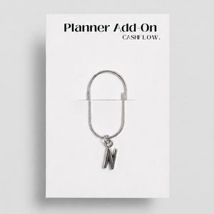 Könnte beinhalten: Eine silberfarbene Büroklammer mit einem Anhänger in Form des Buchstabens "N". Die Büroklammer ist an einer weißen Karte mit dem Text "Planner Add-On" und "CASHFLOW." befestigt.