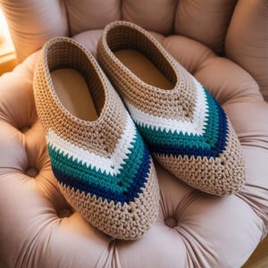 May include: A pair of handmade crochet slippers in a neutral beige color. The slippers feature a chevron design in white, teal, and navy blue. The interior of the slippers is a light tan color. The slippers are resting on a pink upholstered chair.