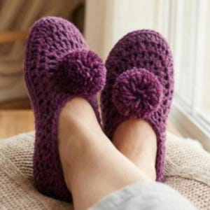 Peut inclure: Paire de pantoufles en crochet violet faites à la main, avec un détail pompon sur le dessus. Les pantoufles sont présentées sur les pieds d'une personne, reposant sur une surface de couleur neutre. Les pantoufles semblent douces et confortables.