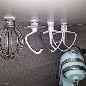 Könnte beinhalten: Eine Sammlung von KitchenAid-Mixer-Aufsätzen, darunter ein Schneebesen, ein Knethaken und ein Flachrührer, die unter einem Schrank montiert sind. Ein hellblauer KitchenAid-Mixer ist darunter zu sehen. Die Aufsätze sind weiß und silber.