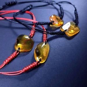 May include: Four amber stone bracelets with adjustable black and red cords. The amber stones vary in shape and have internal patterns. The bracelets are displayed against a dark blue background.