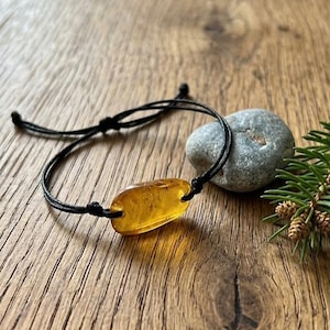 Peut inclure: Un bracelet artisanal avec une pierre d'ambre polie, fixée par un cordon noir. L'ambre a une teinte jaune doré chaleureuse. Le bracelet est présenté sur une surface en bois, avec une pierre grise et une brindille de verdure en arrière-plan.