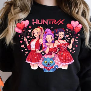 Może przedstawiać: Czarna bluza z grafiką przedstawiającą trzy kobiety w stylu anime trzymające balony w kształcie serca. Kobiety ubrane są w czerwone stroje i otoczone konfetti w kształcie serca. Słowo "HUNTRAX" jest wydrukowane na różowo.