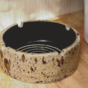May include: A round ceramic ashtray featuring a speckled beige exterior with brown accents and a smooth, solid black interior. The design includes three evenly spaced notches along the top rim for resting items, with a circular grooved pattern at the base.