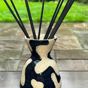 May include: A small ceramic vase with a flared neck and a wide base, featuring a speckled cream and black abstract pattern. Several thin, black incense sticks are inserted into the opening of the vase, extending upwards from the centre.