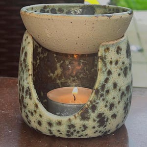 May include: A ceramic oil burner with a speckled light green and dark brown glaze. The structure features a bowl at the top for wax or oil and a hollowed-out base with an arched opening containing a small, lit tea light candle inside.