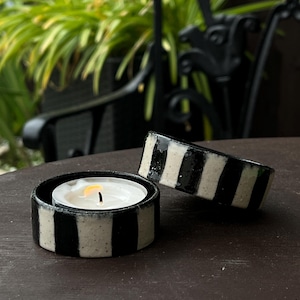 May include: Two circular ceramic candle holders featuring a bold black and white vertical striped pattern. One holder contains a small white tea light candle with a lit flame, while the other rests tilted on its side next to it.