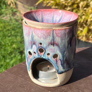 May include: A cylindrical ceramic wax warmer featuring a reactive glaze in shades of pink, blue, and purple. The top section is a removable bowl, while the base has a large arched opening containing a lit tea light candle and small circular vents.