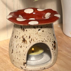 May include: A ceramic oil burner shaped like a mushroom with a glossy red cap featuring white spots. The base is a speckled cream colour with a wide arched opening at the front containing a small, lit yellow tea light candle inside.