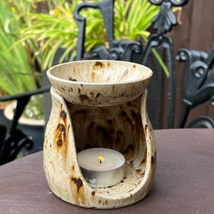 May include: A ceramic oil burner with a speckled cream and brown glaze. The structure features a wide base with an arched opening for a tea light candle and a removable bowl on top. A small white tea light candle with a lit flame sits inside.