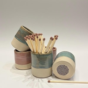 May include: A collection of cylindrical ceramic match holders with a two-tone glaze in shades of blue, pink, and cream. One holder is positioned horizontally to reveal a circular strike pad on its base. A cluster of wooden matches is inside one holder.
