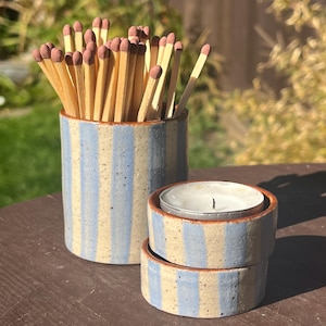 May include: A cylindrical ceramic match holder with vertical light blue stripes on a speckled cream background, filled with wooden matches with brown tips, alongside a matching small ceramic tea light candle holder with a white candle.