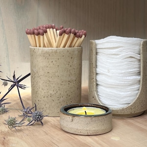 May include: A speckled beige ceramic matchstick holder filled with wooden matches, a matching ceramic cotton pad dispenser containing white round pads, and a small low-profile ceramic candle holder with a yellow tea light candle.