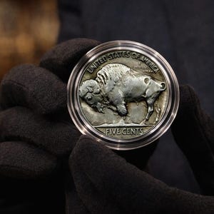 May include: A silver coin showing a bison, with the inscriptions "UNITED STATES OF AMERICA" and "FIVE CENTS". The coin is in a clear protective case, held by gloved hands. A numismatic item.