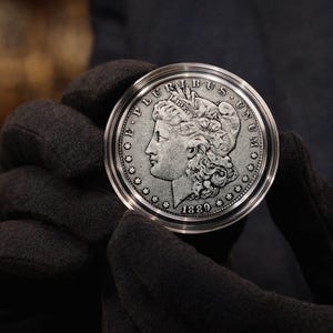 Puede incluir: Primer plano de una moneda de plata Morgan de 1889, dentro de una funda protectora transparente. La moneda presenta un perfil de mujer y las palabras "E PLURIBUS UNUM" y "LIBERTY". La moneda es sostenida por una persona con guantes negros.