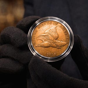 Puede incluir: Una moneda dorada de 1858, dentro de un soporte protector transparente. La moneda presenta un diseño de águila y las palabras "UNITED STATES OF AMERICA". La moneda es sostenida por una persona con guantes negros.