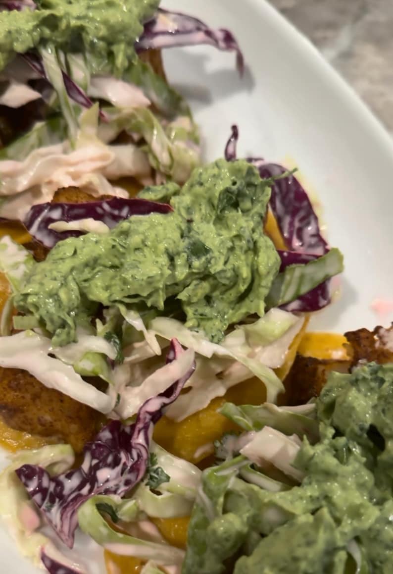 Op de afbeelding: Close-up van een bord met een kleurrijk gerecht. Het gerecht heeft een basis van goudbruin voedsel, gegarneerd met een romige groene saus, geraspte witte en paarse kool en een romige witte saus. Het eten is op een wit bord gerangschikt.