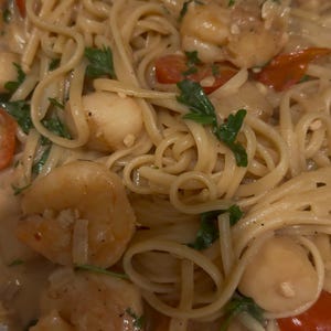 Op de afbeelding: Close-up van een pasta gerecht met zeevruchten. Linguine-noedels worden gemengd met garnalen, sint-jakobsschelpen en gehalveerde kerstomaatjes in een romige saus. Verse peterselie voegt een vleugje groen toe. Het gerecht is klaar om te eten.