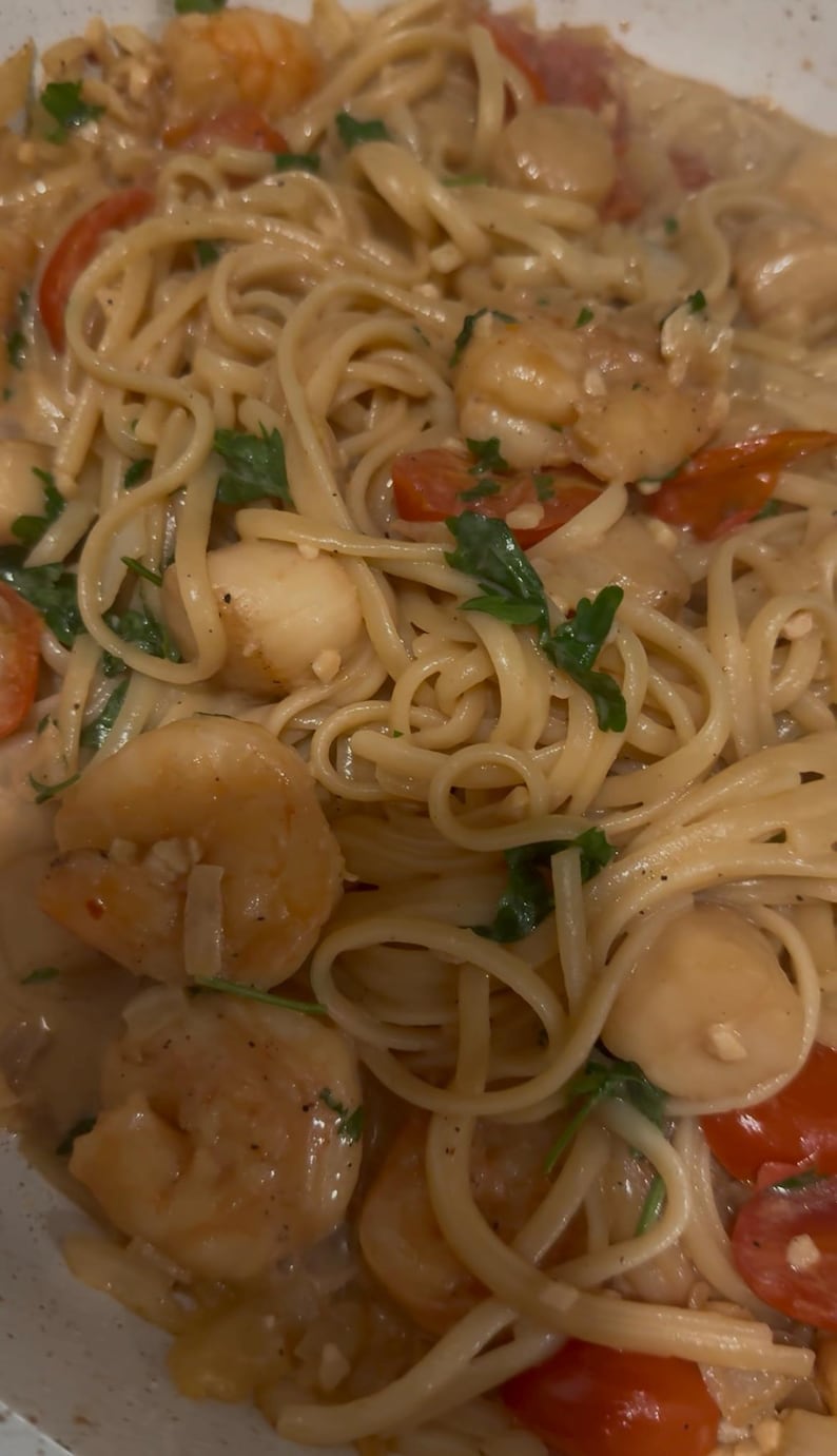 Op de afbeelding: Close-up van een pasta gerecht met zeevruchten. Linguine-noedels worden gemengd met garnalen, sint-jakobsschelpen en gehalveerde kerstomaatjes in een romige saus. Verse peterselie voegt een vleugje groen toe. Het gerecht is klaar om te eten.