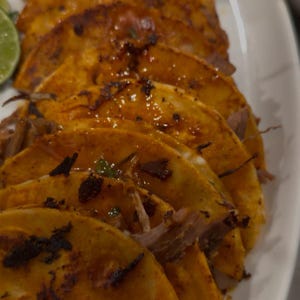 Op de afbeelding: Close-up van verschillende goudbruine taco's op een wit bord. De taco's hebben een knapperige buitenkant en zijn gevuld met vlees en gegarneerd met groene kruiden. Een limoenpartje is zichtbaar in de hoek.