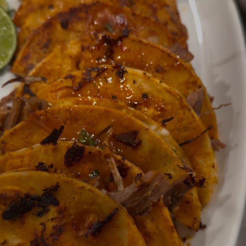 Op de afbeelding: Close-up van verschillende goudbruine taco's op een wit bord. De taco's hebben een knapperige buitenkant en zijn gevuld met vlees en gegarneerd met groene kruiden. Een limoenpartje is zichtbaar in de hoek.