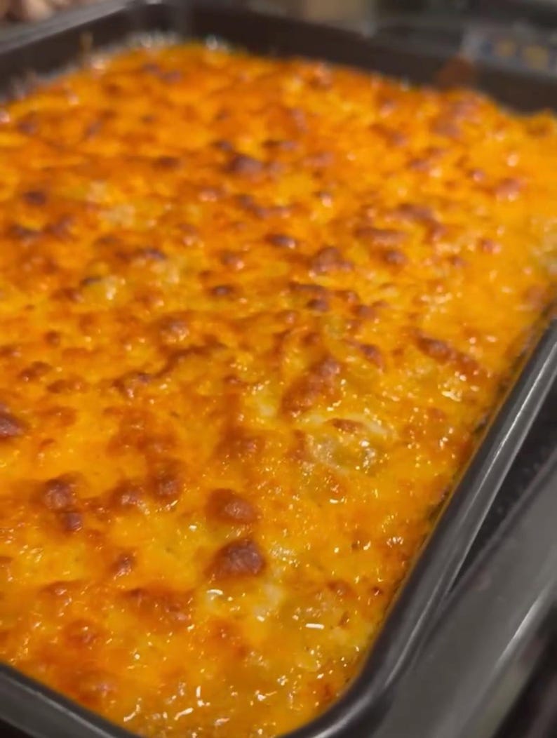 Op de afbeelding: Een close-up van een gebakken ovenschotel met macaroni en kaas. De bovenkant is goudbruin en borrelt, met een rijke, kazige textuur. De schotel bevindt zich in een donkere, rechthoekige bakvorm.