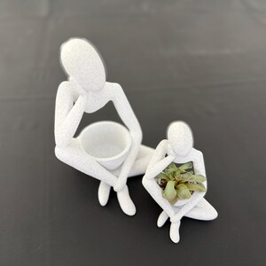 May include: Two white, abstract figure planters. One figure is seated with a large bowl, the other holds a small succulent plant. The planters have a textured surface and are on a dark background.