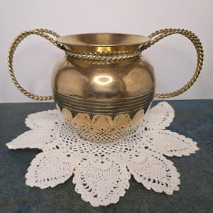 May include: A vintage brass vase with two twisted rope handles. The vase has a rounded body with decorative bands and sits on a white, crocheted doily. The vase is a warm gold color.