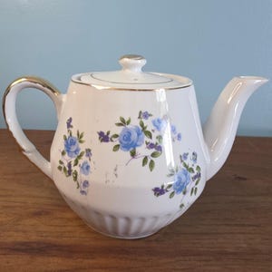 Op de afbeelding: Witte keramische theepot met een gouden handvat en rand, versierd met blauwe rozenbloempatronen en paarse accenten. De theepot heeft een deksel met een gouden knop en een gebogen tuit. Een vintage theeservies item.
