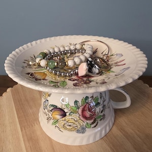 May include: A decorative jewelry holder made from a repurposed teacup and saucer. The white ceramic piece features a floral pattern with pink, yellow, and purple flowers. Several bracelets and rings rest on the saucer, including beaded and metal designs.