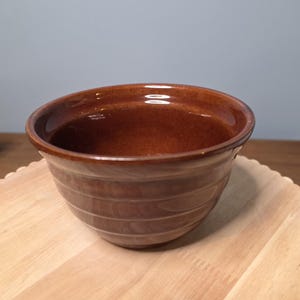 Könnte beinhalten: Eine braune Keramikschüssel mit glänzendem Finish. Die Schüssel hat eine abgerundete Form mit horizontalen Rillen an der Außenseite. Sie steht auf einer hellen Holzoberfläche.