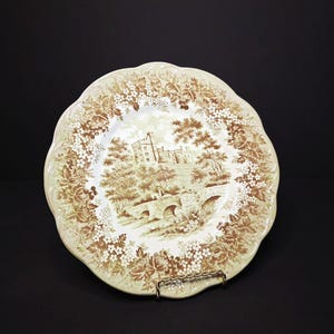 Puede incluir: Un plato decorativo con borde festoneado, que presenta un paisaje marrón y blanco de un castillo, un puente y árboles. El borde está adornado con motivos florales. El plato se exhibe sobre un soporte metálico sobre un fondo negro.