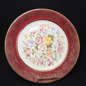 May include: A decorative plate with a red border and a gold rim. The center of the plate features a floral design with pink, yellow, and blue flowers. The red border has a gold filigree pattern.