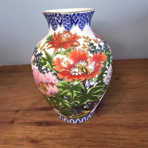 May include: A white ceramic vase with a floral design. The vase features a blue patterned rim and base, with a gold trim at the top. The body of the vase is decorated with large orange, pink, and white flowers, surrounded by green foliage.